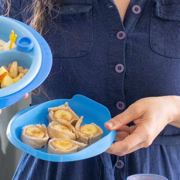 Mae_segurando_pratos_comida Mãe segurando dois pratos de comida azuis. No lado esquerdo, há um prato com duas divisórias contendo tomate cereja, azeitona, queijo, uva, maçã e amendoas. No prato mais á direita, há um "sushi" de banana (banana enrolada em um pão sírio).