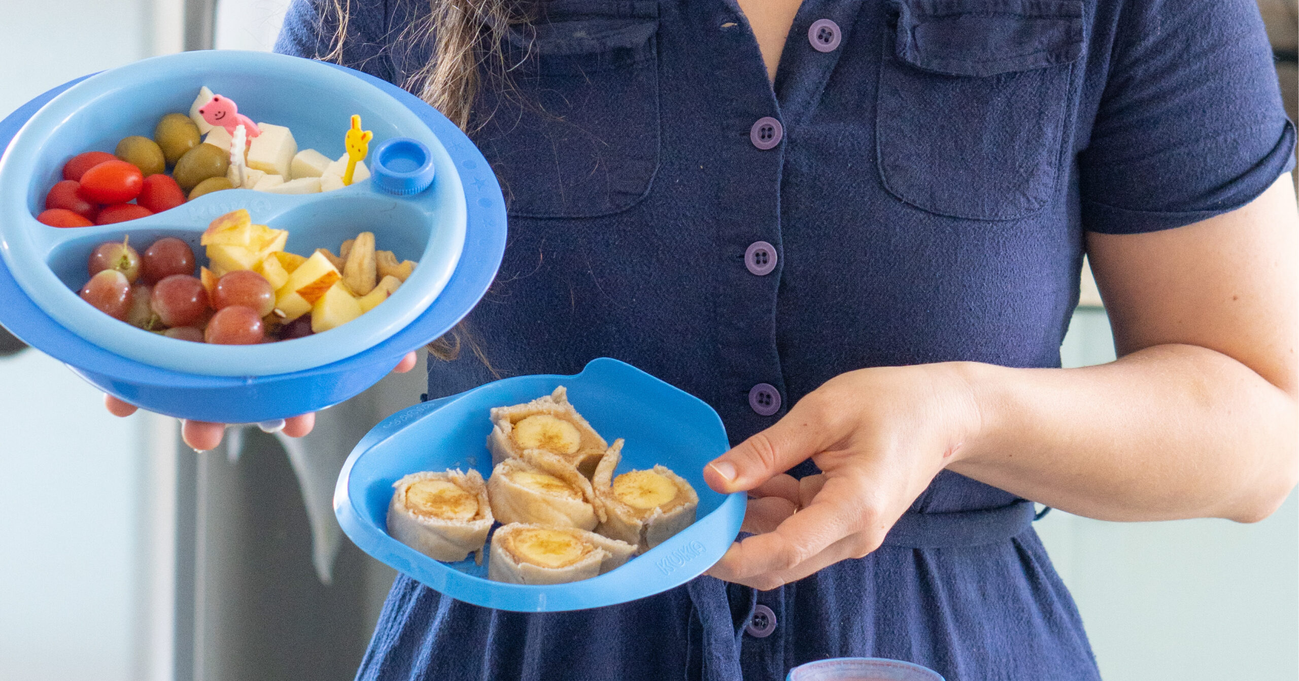 Mãe segurando dois pratos de comida azuis. No lado esquerdo, há um prato com duas divisórias contendo tomate cereja, azeitona, queijo, uva, maçã e amendoas. No prato mais á direita, há um "sushi" de banana (banana enrolada em um pão sírio).
