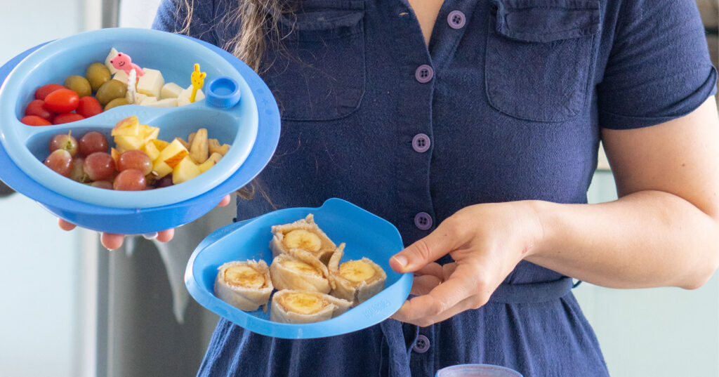 Mãe segurando dois pratos de comida azuis. No lado esquerdo, há um prato com duas divisórias contendo tomate cereja, azeitona, queijo, uva, maçã e amendoas. No prato mais á direita, há um "sushi" de banana (banana enrolada em um pão sírio).
